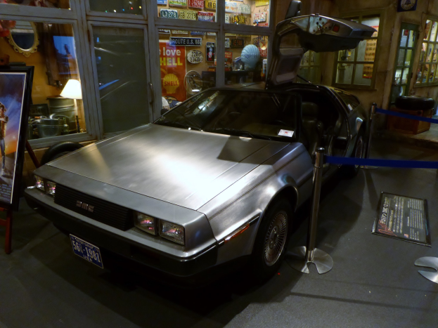 Ein DeLorean DMC-12 aus der Back to the Future-Reihe wird in einem Museum ausgestellt, dessen Türen offen sind und umgeben von Informationsanzeigen, Bändern und verschiedenen Gegenständen.