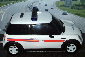 Weißes Polizeiauto vor einer belebten Straße mit Bäumen und Gras auf beiden Seiten, aufgenommen ausserhalb der Stadt.