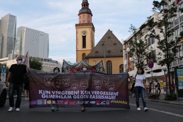 Eine Gruppe von Menschen in Masken, die eine Straße entlanggehen und ein Banner halten, mit einem geparkten Auto auf der linken Seite, Gebäuden, Bäumen, Schildern, Polen, einem Uhrenturm und einem klaren blauen Himmel im Hintergrund, was auf eine Protestaktion gegen Rassismus hindeutet.