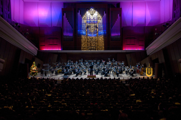 Ein großes Auditorium voller Menschen bei einem Konzert, mit Musikern auf der Bühne und anderen im Publikum, einem Weihnachtsbaum im Hintergrund und Wänden mit Lichtern und anderen Gegenständen geschmückt.