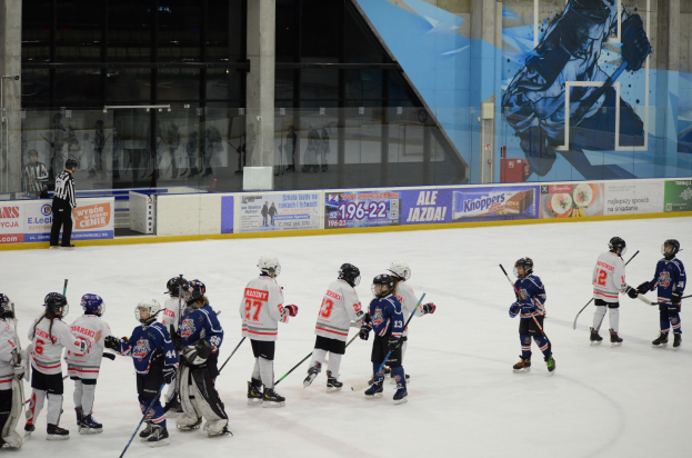 Junge Menschen in Helmen und Eishockey-Ausrüstung spielen Eishockey auf einer Indoor-Eisbahn, mit Bannern, Säulen und Glaswänden im Hintergrund.
