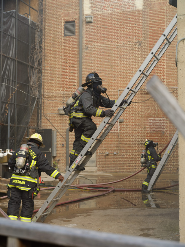 Feuerwehrleute in Helmen und Ausrüstung klettern an einer Leiter vor einem Backsteingebäude mit Rohren und einem Metallstab im Vordergrund, mit einem weiteren Gebäude im Hintergrund.