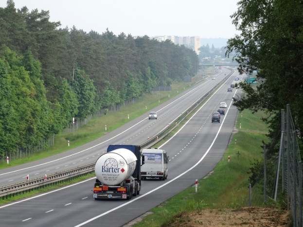 Ein großer Lastwagen fährt auf einer von Bäumen gesäumten Autobahn, mit einem Zaun und Gras auf der rechten Seite und Gebäuden in der Ferne unter einem klaren blauen Himmel.