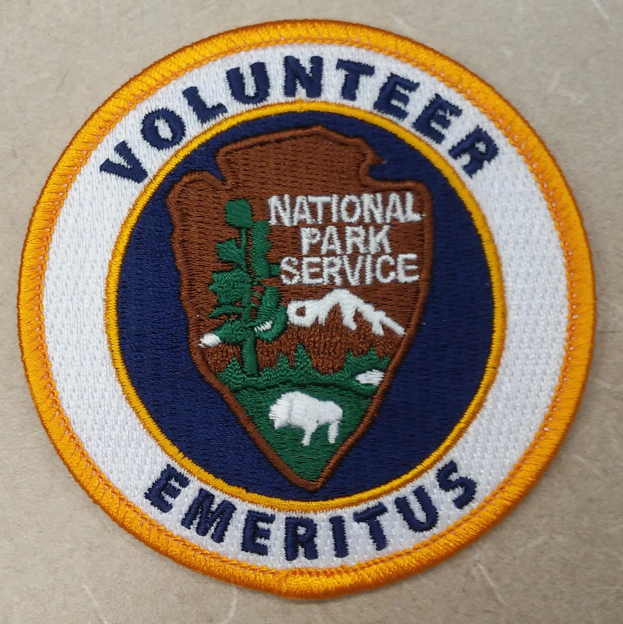Ein kreisförmiges Patch mit einem blauen Rand, einem weißen Hintergrund und fettem schwarzem Text, der "Volunteer National Park Service Emeritus" in der Mitte eines gelben Kreises liest.