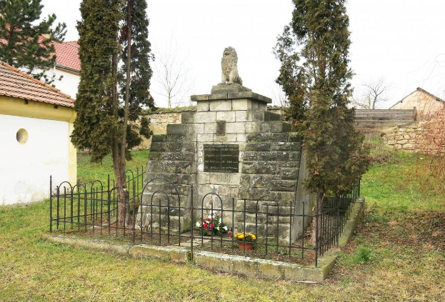 Ein steinernes Holocaust-Gedenkmal in einer grünen Wiese, umgeben von einem Metallzaun und geschmückt mit Blumensträußen, mit Bäumen, Häusern und einer Wand im Hintergrund unter einem klaren blauen Himmel, gelegen in Sibiu, Rumänien.