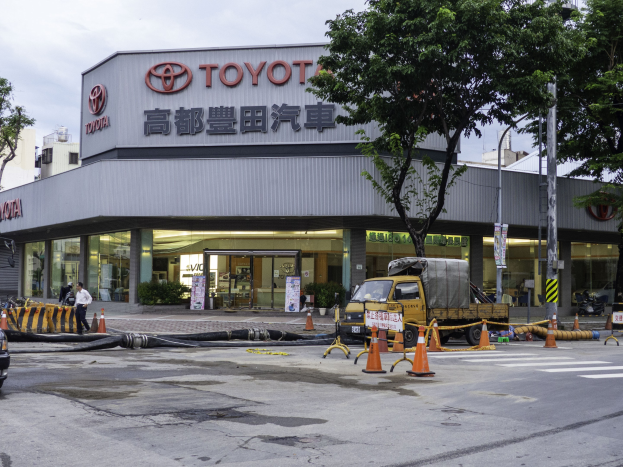 Ein Toyota Autohaus in Tokio, Japan, mit Fahrzeugen, Verkehrskegeln, Rohren, Menschen, Bäumen, Polen, Brettern und Gebäuden im Vordergrund und einem bewölkten Himmel im Hintergrund.