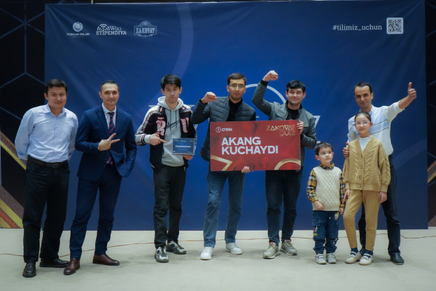 Eine Gruppe von Menschen auf einer Bühne, die eine Tafel mit der Aufschrift 'Akang Kuchaydi - Zakovat Quiz' halten, während im Hintergrund ein Banner mit Text und einem Barcode zu sehen ist.