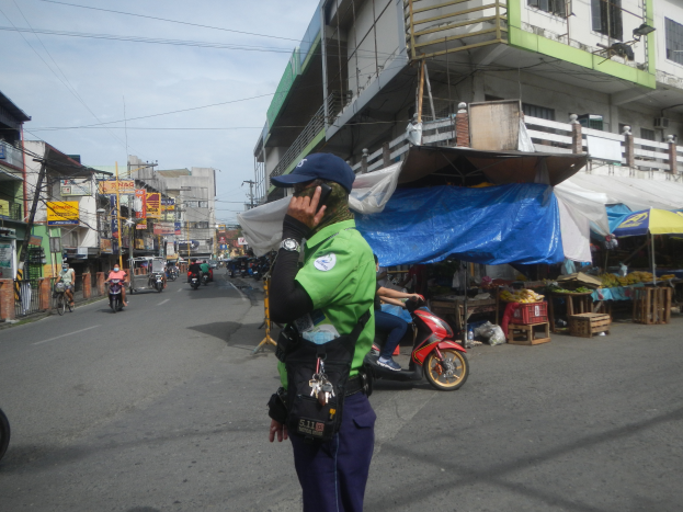 Ein Polizist in einer grün-blauen Uniform und einer blauen Mütze steht auf der Seite einer Straße und telefoniert mit einem Handy, während im Hintergrund Fahrzeuge fahren, rechts von ihm Stände mit Gegenständen und ein bewölkter Himmel.