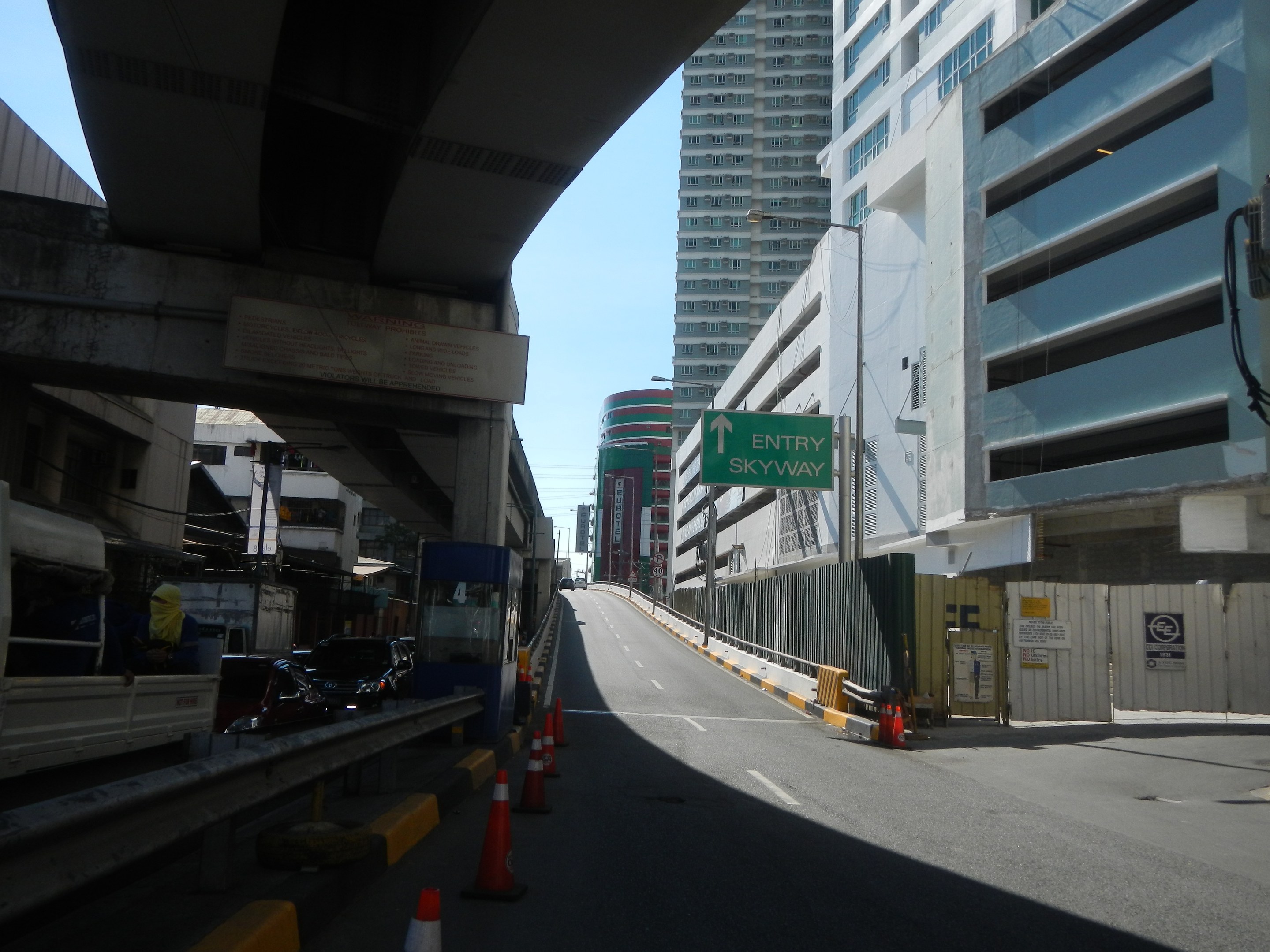 Stadtstraße mit Verkehrskegeln, Fahrzeugen, einer Brücke auf der linken Seite, Gebäuden mit Namensschildern, Laternenmästen und einem Himmel im Hintergrund.