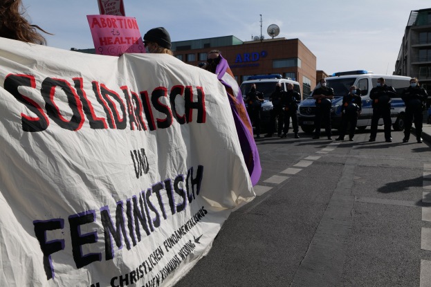 Gruppe von Menschen marschiert auf einer Straße mit einem "Solidarität und Feminismus"-Schild, geparkte Fahrzeuge, Gebäude, eine Schüsselantenne und einen klaren blauen Himmel im Hintergrund.