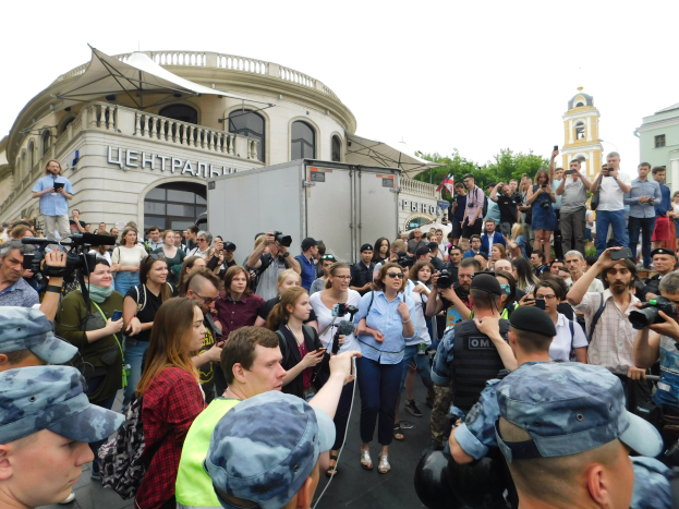 Große Gruppe von Menschen demonstriert vor einem Gebäude, einige halten Kameras und Handys, mit einem Fahrzeug, Bäumen und einem klaren blauen Himmel im Hintergrund.