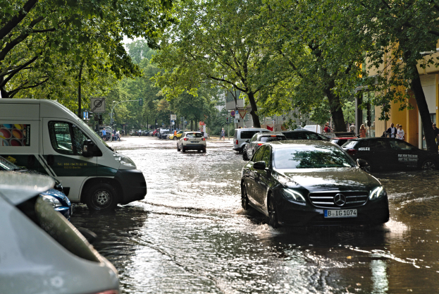 Eine überflutete Straße in Berlin, Deutschland, mit Autos, die durch das Wasser fahren, Menschen, die auf dem Gehweg gehen, Bäume, die die Straße säumen, Gebäude im Hintergrund und Schilder und Pfosten im Wasser.