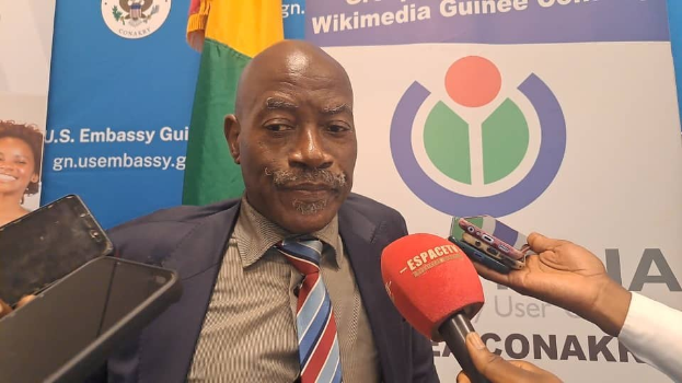 Ein Mann im Anzug spricht mit Reportern, während im Vordergrund eine Person mit einem Mikrofon und einem Gerät zu sehen ist. Im Hintergrund sind Banner, Flaggen und eine Frau zu erkennen.