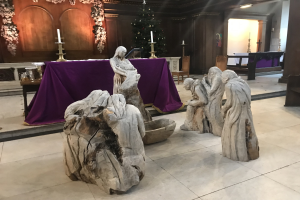 Krippe in einer Kirche mit Statuen, einem Tisch mit einem Tuch und Kerzen, Stühlen, einem geschmückten Weihnachtsbaum und einer Wand im Hintergrund.