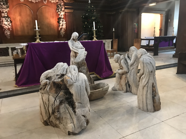 Krippe in einer Kirche mit Statuen, einem Tisch mit einem Tuch und Kerzen, Stühlen, einem geschmückten Weihnachtsbaum und einer Wand im Hintergrund.
