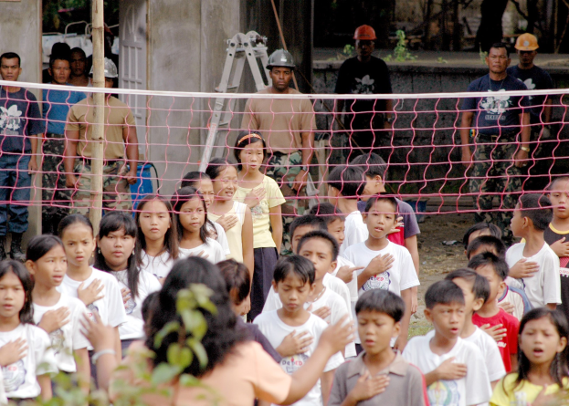 Gruppe von Kindern vor einem Netz mit einer Frau im Vordergrund, Hintergrund enthält Helme tragende Personen, eine Leiter, eine Wand und Bäume.
