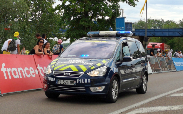 Polizeiauto fährt an einer Menschenmenge mit Schildern, Geländern, Bäumen, einer Brücke, einer Fahne und einem bewölkten Himmel vorbei.