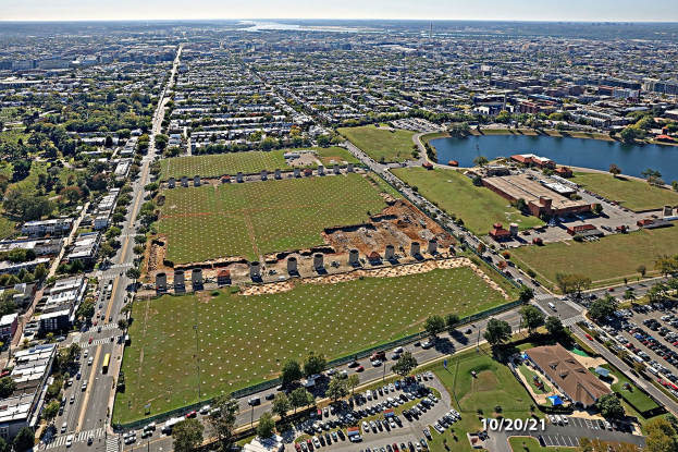 Ein Park mit grünem Gras, Bäumen, Pflanzen, Laternenmasten und Skulpturen vor dem Hintergrund von Gebäuden und einem klaren blauen Himmel.