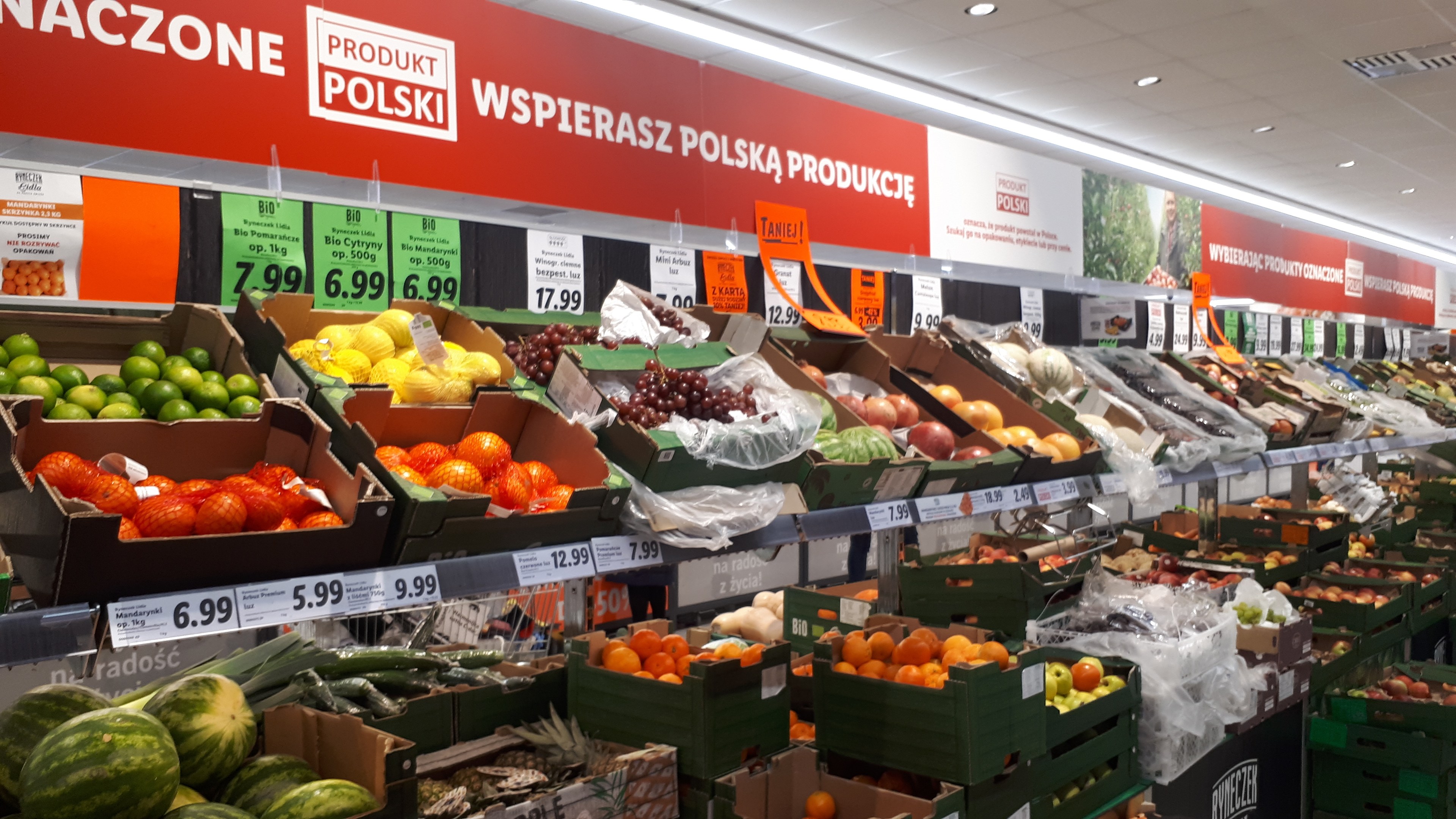 Ein Geschäft mit verschiedenen Obst- und Gemüsesorten in Kartons mit Preisschildern, Informationsschilder an den Wänden und Deckenleuchten.