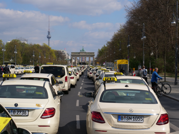Eine lange Reihe von Taxis steht entlang einer belebten Straße in Berlin, Deutschland, mit Fahrradfahrern und Fußgängern auf dem Gehweg, flankiert von Bäumen und Laternenmasten und Gebäuden, einem Bogen und einem Turm im Hintergrund unter einem bewölkten Himmel.