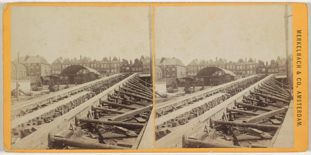 Ein altes Schwarz-Weiß-Foto einer Bahnschiene mit Gebäuden im Hintergrund, Text auf der rechten Seite mit 'Merkelbach & Co. Amsterdam' und ein paar hölzerne Gegenstände im Vordergrund.