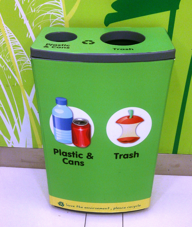 Ein grüner Mülltonne mit Plastik und Dosen auf einem weißen Boden, mit einem Plakat an der Wand im Hintergrund.