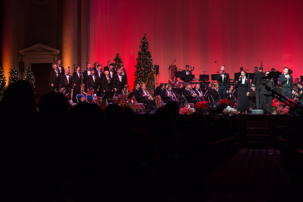 Eine Gruppe von Menschen, die auf der Bühne mit Musikinstrumenten spielt, umgeben von Blumenbouquets, vor geschmückten Weihnachtsbäumen und Säulen unter einem roten Vorhang.