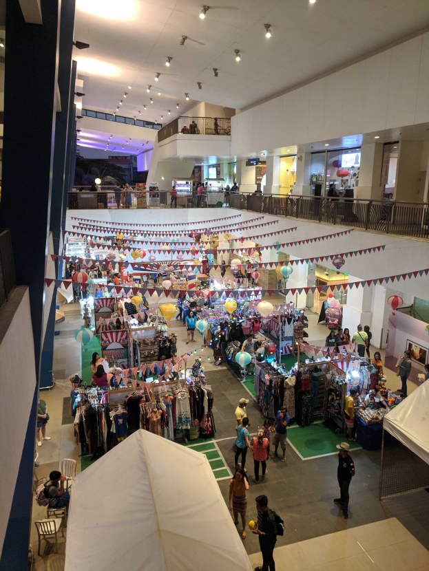Eine feierliche Menge schlendert durch einen belebten Indoor-Markt mit Kleidungsständen, dekorativen Fahnen, Luftballons und Sitzbereichen, wahrscheinlich auf einem Weihnachtsmarkt im Mall of Asia.