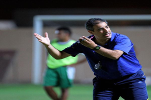 Ein Mann in einem blauen T-Shirt und Hose steht auf einem Fuballfeld und hält ein Mikrofon in der Hand, wobei er auf Spieler zeigt. Im Hintergrund sind eine Person in einem grünen Kleid, ein Tor und eine Wand zu sehen.