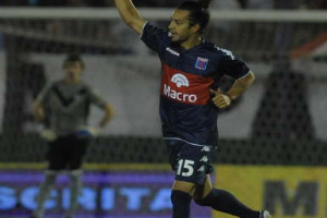 Ein Fussballer feiert nach einem Tor auf dem Boden, mit jubelnden Zuschauern und einer Person im Hintergrund und einer Tafel mit Text dahinter, alles vor einem leicht unscharfen, energetischen Hintergrund.