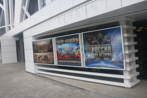 Großes Plakat an einem Gebäude, das die Eröffnungszeremonie der Olympischen Spiele in Moskau ankündigt, mit beleuchtetem Text und Bildern und einem Teil des Bodens unten sichtbar.