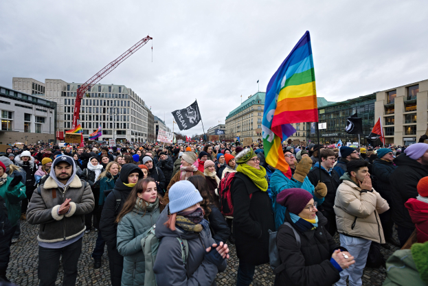 Eine große Gruppe von Menschen mit Fahnen und Transparenten vor einem Gebäude während einer LGBTQ+-Rechtsdemo in Berlin, mit Gebäuden, einem Kran und Wolken im Hintergrund.