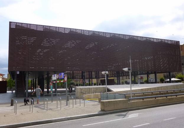 Großes modernes Gebäude mit zahlreichen Fenstern, das als neuer Eingang zur City dient, umgeben von Straßeninfrastruktur, Fahrzeugen, Fußgängern, Bäumen und einem bewölkten Himmel.