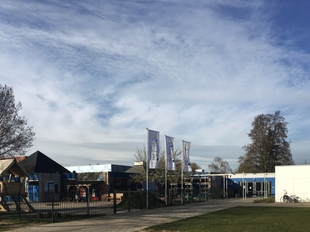 Großes Schulgebäude mit Metallzaun, Rasenfläche, Weg, Bäumen, Bannern, einem geparkten Fahrrad, nahen Häusern und einem bewölkten Himmel, mit einem neuen Spielplatz für Kinder.