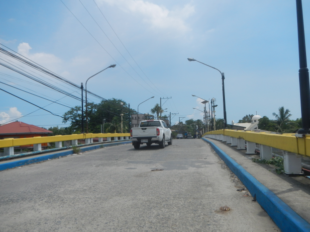 Ein Lkw fährt über eine Brücke mit gelben Geländern, umgeben von Laternenmasten, Strommasten mit Drähten, Bäumen, Gebäuden und einem bewölkten Himmel.