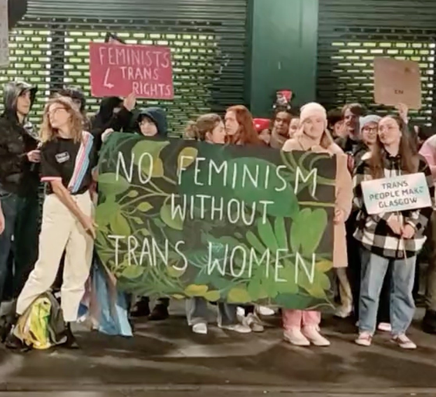Eine Gruppe von Menschen hält ein Transparent mit der Aufschrift "Kein Feminismus ohne Transfrauen" und Schilder in der Öffentlichkeit.
