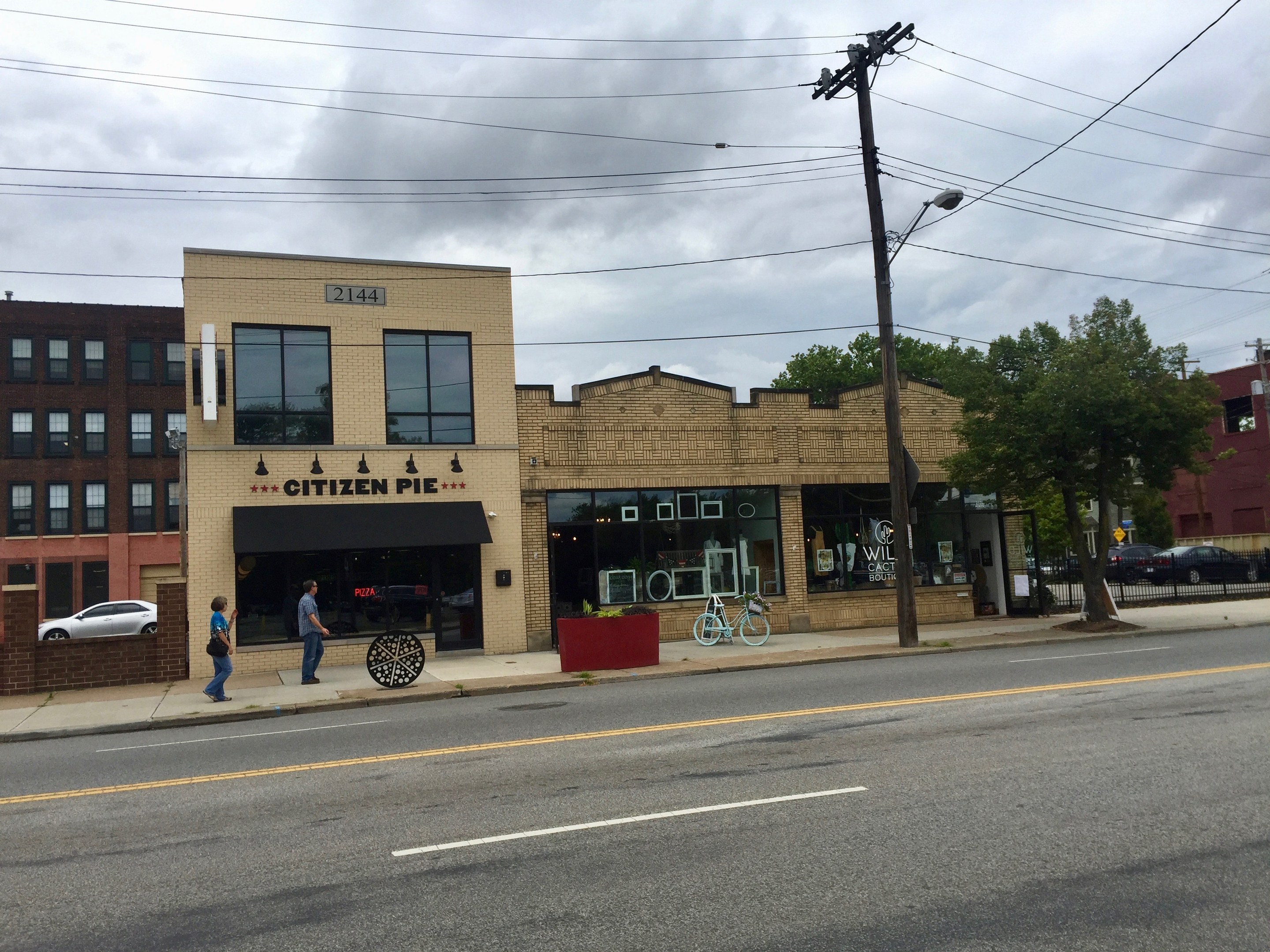 Eine belebte Stadtstraße mit Fußgängern, parkenden Fahrrädern, Strommasten, Bäumen und Gebäuden, mit dem Citizen Pie Restaurant an der Ecke unter einem bewölkten Himmel.