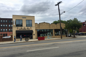 Eine belebte Stadtstraße mit Fußgängern, parkenden Fahrrädern, Strommasten, Bäumen und Gebäuden, mit dem Citizen Pie Restaurant an der Ecke unter einem bewölkten Himmel.