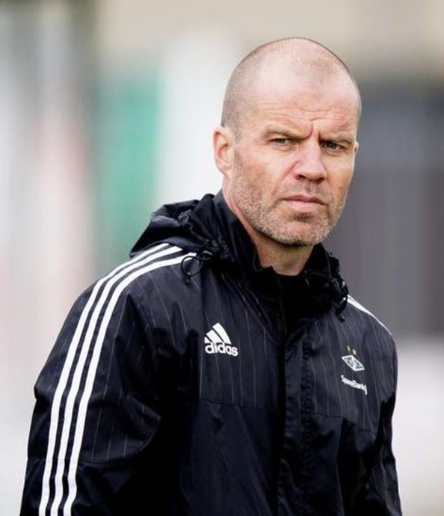 Ein kahlköpfiger Trainer in einer blauen Jacke mit weißen Streifen steht auf einem Fußballfeld, die Hände verschränkt und mit einem ernsten Gesichtsausdruck vor einem unscharfen Hintergrund.