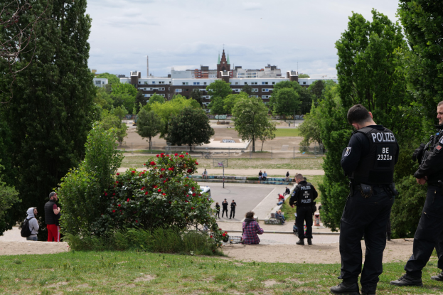 Zwei Polizeibeamte vor einer Gruppe von Menschen in einem Park mit saftig grünem Gras, Bäumen, bunten Blumen, Gebäuden, Pfählen und einem klaren blauen Himmel.