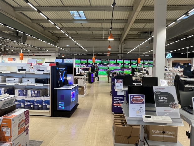 Ein großer Raum in einem Walmart-Laden voller Elektronik, die in Regalen angeordnet ist, einschließlich Computer und Drucker, mit Menschen auf dem Boden und einer Säule, Schildern, Deckenleuchten und Deckenventilatoren.