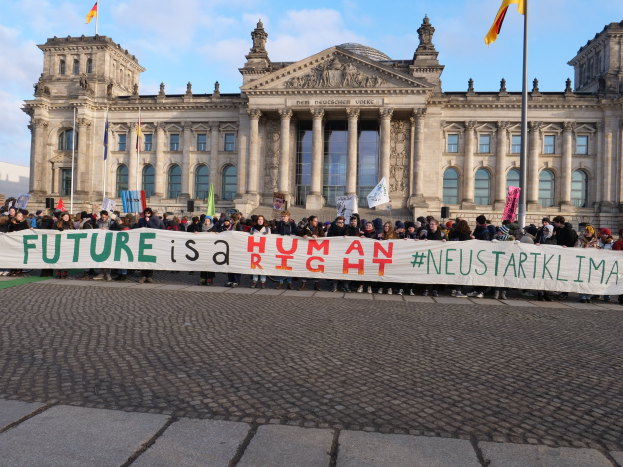 Eine Gruppe von Menschen hält ein Transparent mit der Aufschrift "Zukunft ist ein Mensch" vor dem Reichstaggebäude in Berlin, Deutschland, mit den architektonischen Details des Gebäudes und Fahnen im Hintergrund bei bewölktem Himmel.