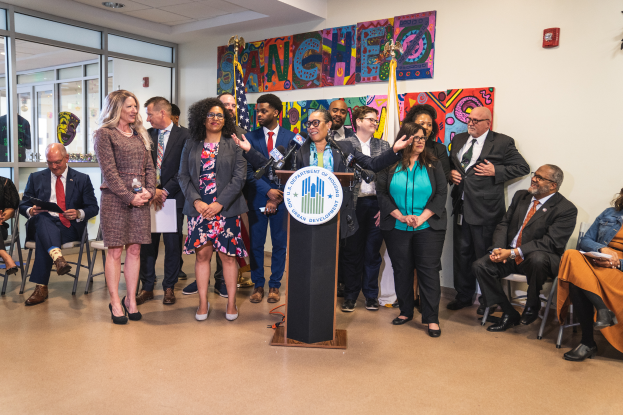 Eine Gruppe von Menschen, die vor einem Podium mit Mikrofon und einer Tafel mit der Aufschrift "Sancho City Councilman Joins City Council" stehen und sitzen, mit Flaggen, Feuerlöscher und Deckenlampen an der Rückwand.