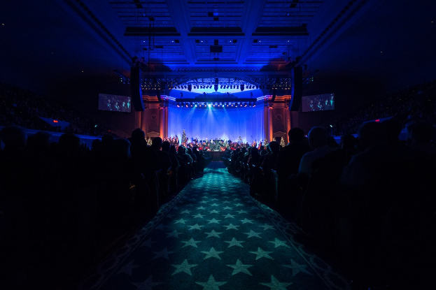 Ein großer, schwach beleuchteter Zuschauerraum mit Sitzreihen, die einer Bühne mit Musikern gegenübersitzen, mit Deckenbeleuchtung und Bildschirmen, was auf einen 2019 Nationalen Kongress-Konzerthalle hindeutet.