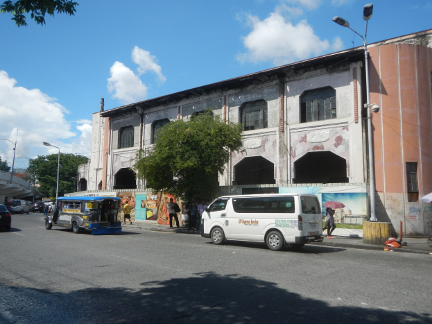 Eine belebte Stadtstraße mit Fahrzeugen, Fußgängern, Bäumen, Laternenmasten, einem großen Gebäude mit Wandgemälde, einer fernen Brücke und einem bewölkten Himmel.