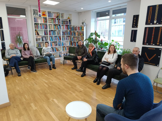 Eine Gruppe von Menschen sitzt um einen Tisch in einem Raum mit Bücherregalen, Pflanzen und Deckenleuchten und ist in ein Gespräch vertieft während eines Workshops zur Gesundheit und Wellness in Berlin, Deutschland.
