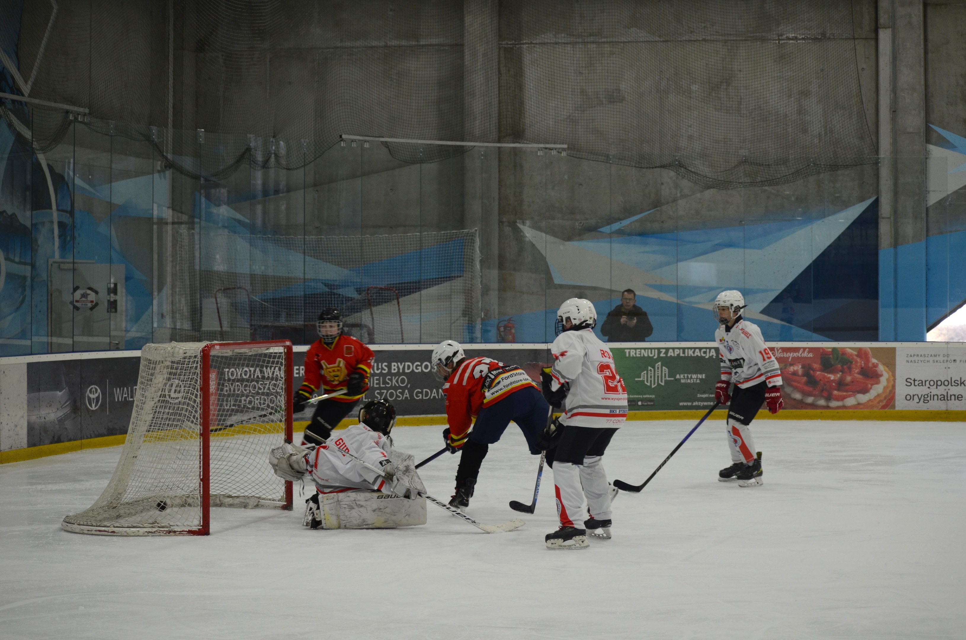 Eine Gruppe von Menschen, die Eis hockey auf einem Eisplatz spielen, tragen Helme und halten Hockey-Schläger in der Hand, mit einem Torpfosten links und Bannern im Hintergrund.