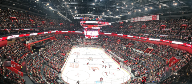 Eine große Eishockeyhalle voller Zuschauer, hell erleuchtet von Deckenlampen, mit einem zentralen Scoreboard und einer enthusiastischen Menge, die während des Spiels jubelt.