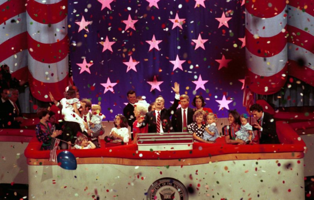 Eine Gruppe von Menschen auf einer Bühne mit einem Rednerpult, das ein Logo, Luftballons und Konfetti im Hintergrund zeigt, mit Sternen und einem Vorhang im Hintergrund.