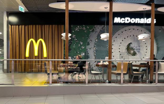 Innenansicht eines McDonald's-Restaurants in einem Einkaufszentrum mit Sitzbereichen, Tischen, einer Glaswand mit Text, Deckenleuchten und sichtbarem Bodenbelag.
